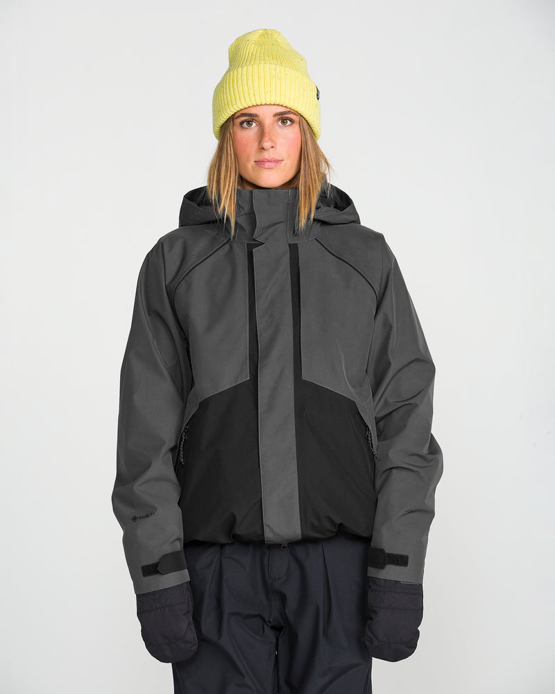 A person wearing a yellow beanie, black gloves, and dark pants stands against a plain white background, sporting the Volcom Women’s Unisex Yourfav Baggy Gore Jacket in gray and black.