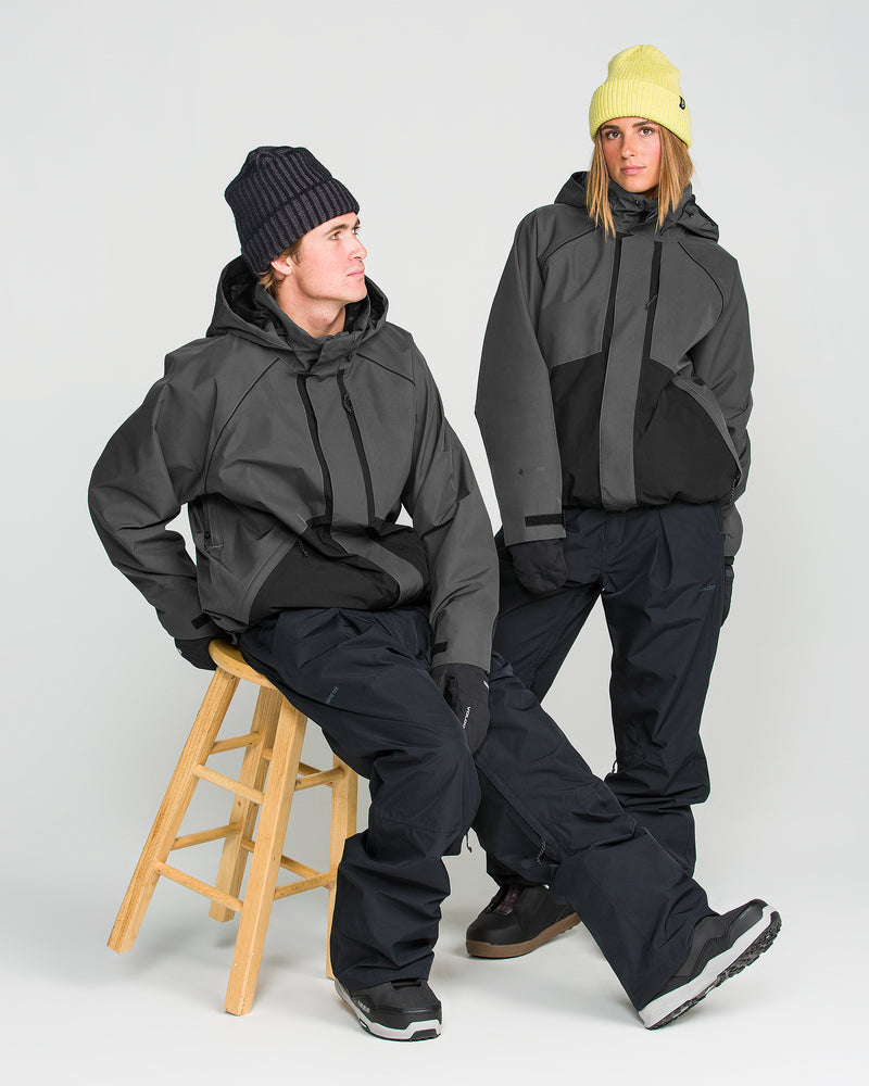 Two people pose against a plain background wearing dark, storm-proof winter outfits. One sits on a wooden stool while the other stands with a hand in their pocket. Both wear chunky boots, pants, and beanies. One of them sports the Volcom Women’s Unisex Yourfav Baggy Gore Jacket—a breathable GORE-TEX jacket—looking stylish and ready for any weather.