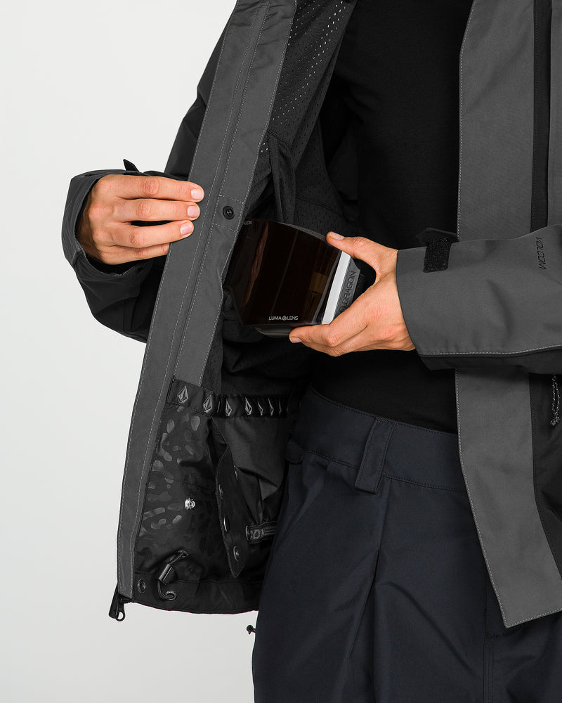 Wearing the Volcom Women’s Unisex Yourfav Baggy Gore Jacket, a person places a rectangular, dark-colored object with a reflective surface into an inner pocket; only their torso and hands are visible.