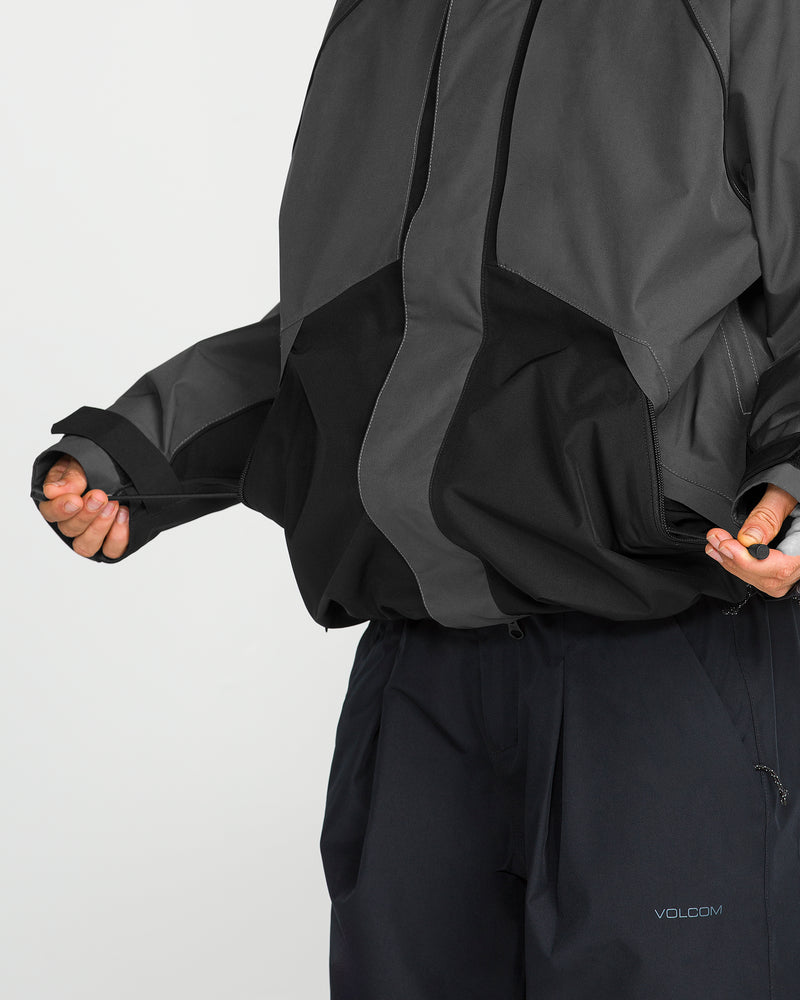 A person pulls at the bottom edge of a dark, oversized Volcom Women’s Unisex Yourfav Baggy Gore Jacket, paired with black pants featuring the VOLCOM logo. The jacket’s breathable, storm-proof design offers comfort in any weather. Their face is not shown.