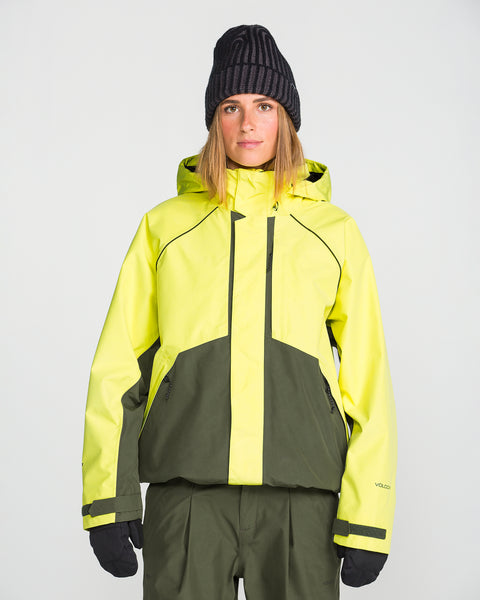 A person wearing the Volcom Unisex Yourfav Baggy Gore Jacket in yellow and green, paired with olive green snow pants, black gloves, and a black knit beanie, standing against a plain white background.