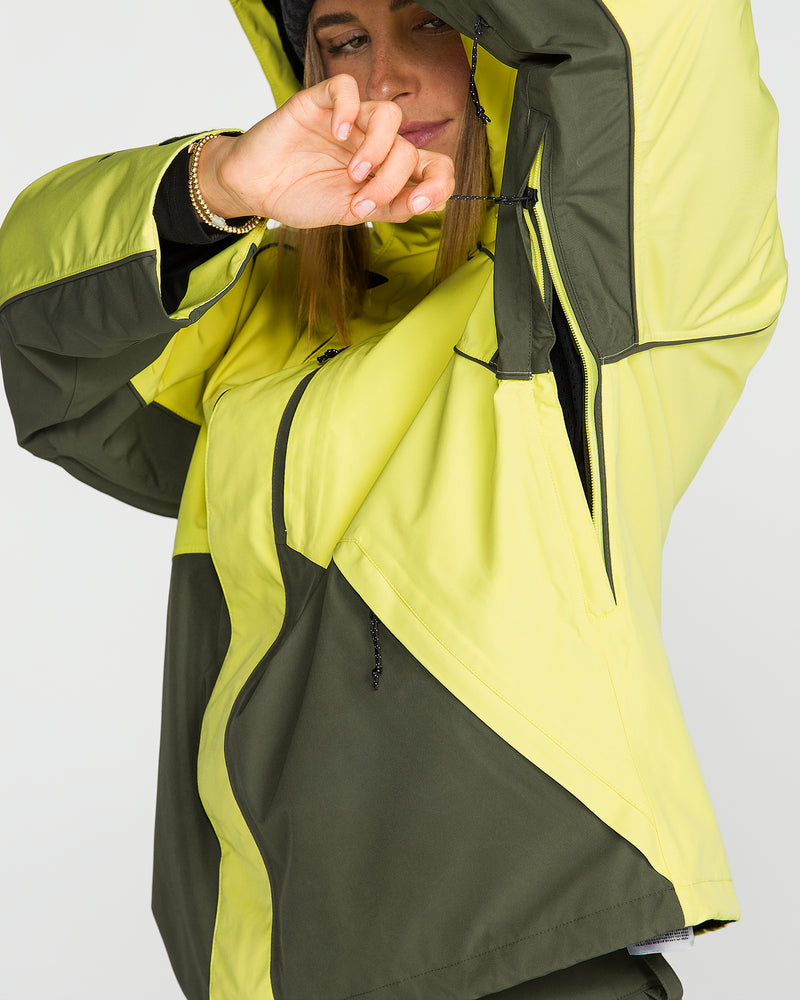 A person wearing the Volcom Unisex Yourfav Baggy Gore Jacket in yellow and olive green pulls the hood over their head while adjusting the front zipper, partially covering their face with one arm.