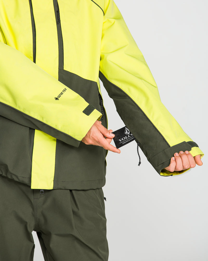 A person wearing a yellow and dark green Volcom Unisex Yourfav Baggy Gore Jacket and matching pants pulls on a black Volcom tag attached to the jacket sleeve. Only the person’s torso and hands are visible. Featuring a GORE-TEX logo on the sleeve, this waterproof Volcom jacket showcases advanced protection against the elements.