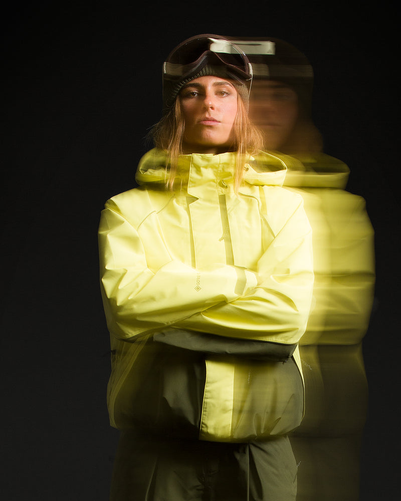 Dressed in the Volcom Unisex Yourfav Baggy Gore Jacket—crafted with storm-proof, breathable GORE-TEX fabric—a person in yellow-green ski gear and goggles stands with arms crossed against a dark background. A motion blur effect in the image creates a dynamic double exposure appearance.