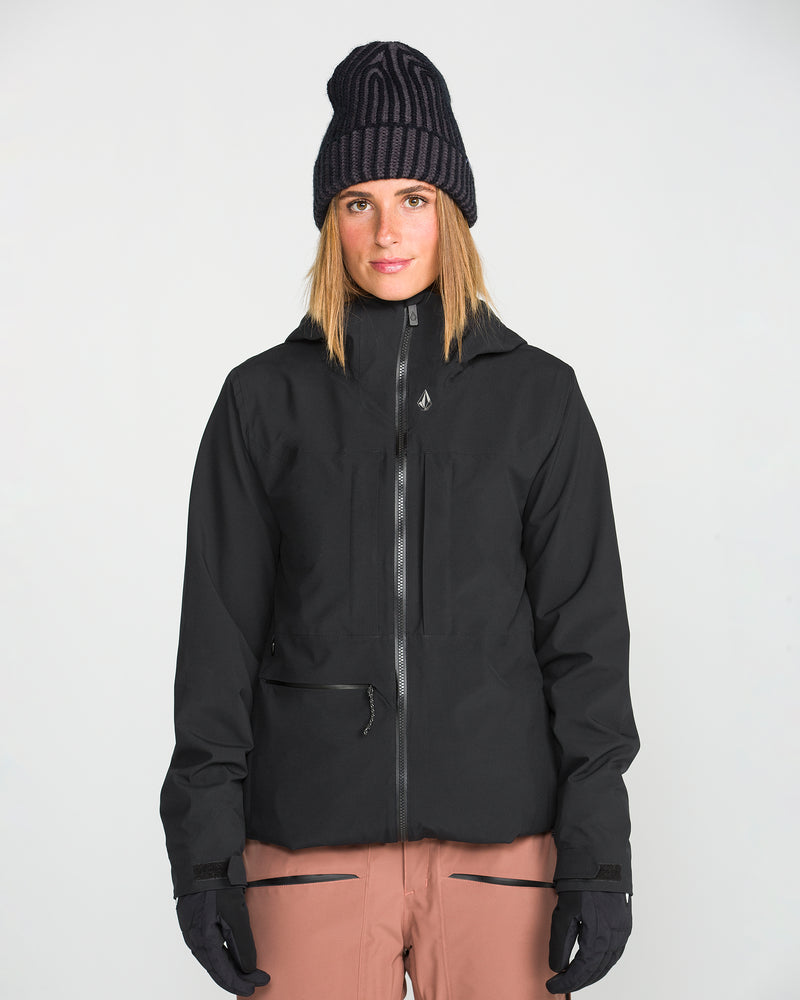 A person wearing a Volcom Women’s Cloudbreaker Stretch 30K Jacket in black with a recycled shell, along with a black knit hat, black gloves, and tan pants, stands facing forward against a plain light background.