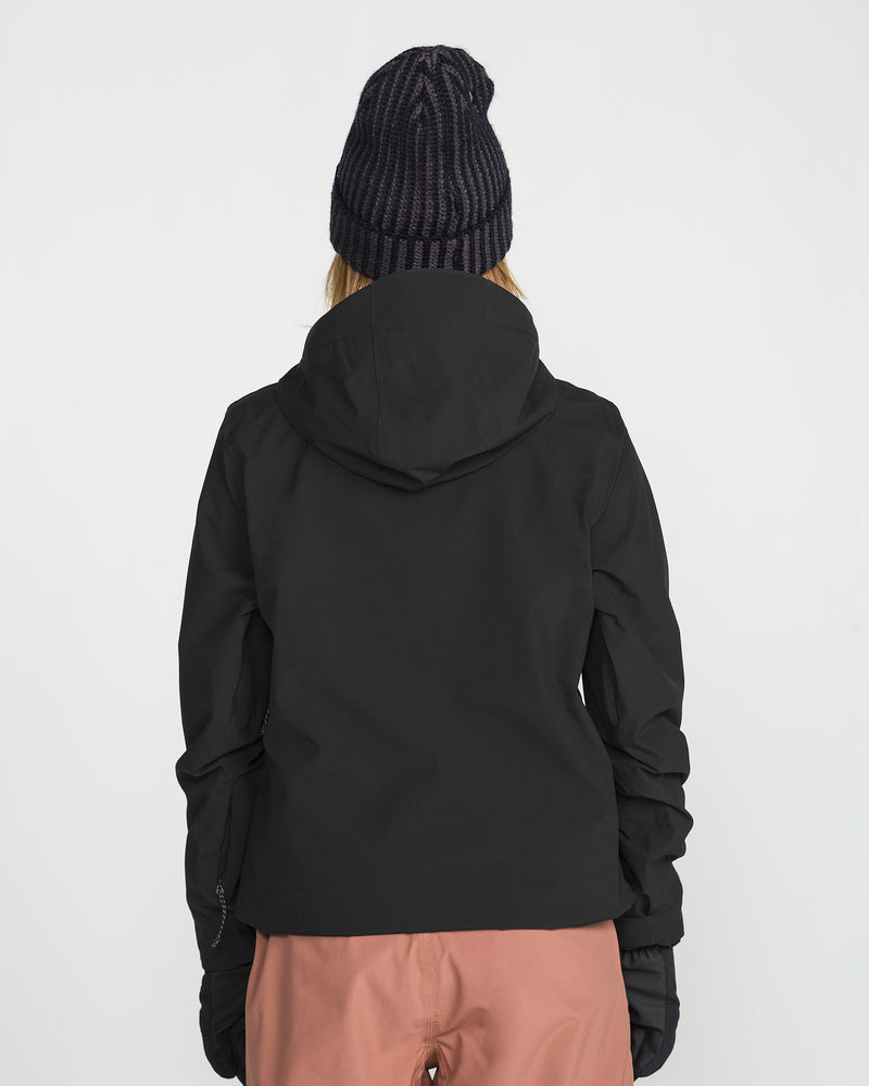 A person is standing with their back to the camera against a plain white background, wearing a black beanie, black gloves, pink pants, and the Volcom Women’s Cloudbreaker Stretch 30K Jacket with a recycled shell.