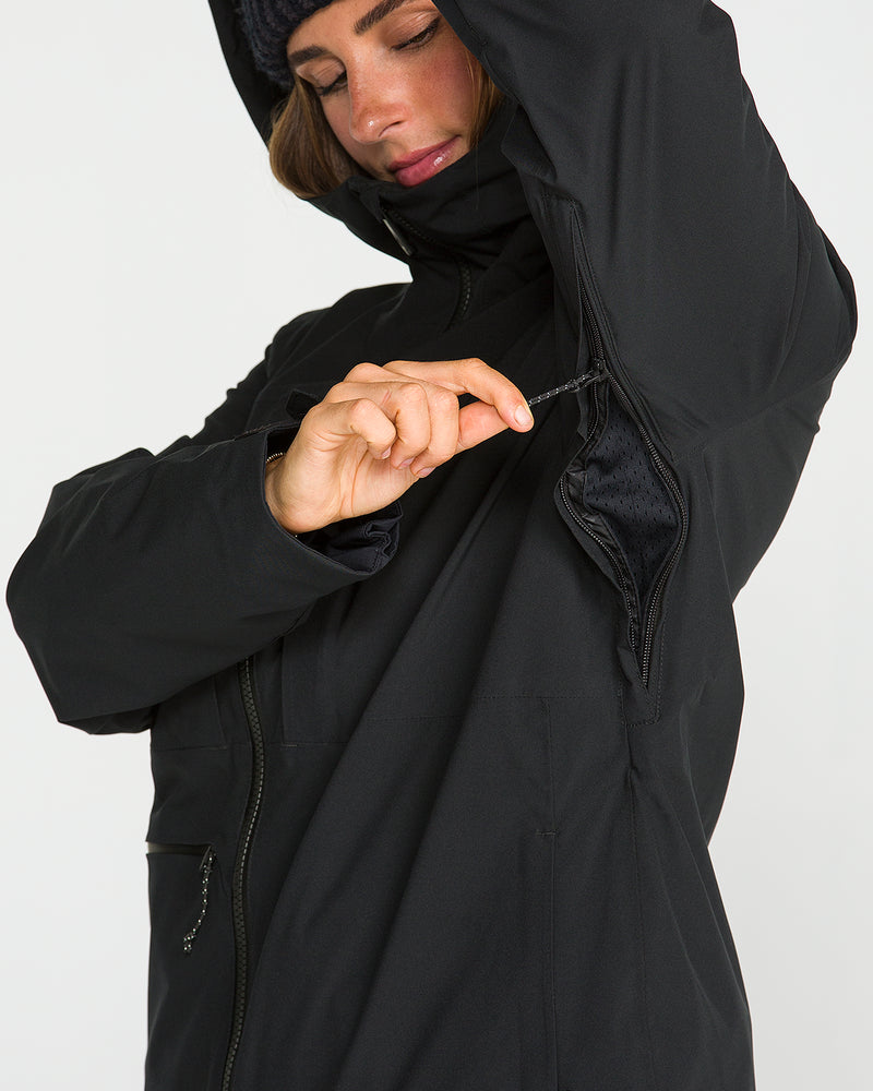 A person wearing the Volcom Women’s Cloudbreaker Stretch 30K Jacket in black and a beanie unzips the underarm ventilation opening. This sleek jacket features a recycled shell and provides 30K waterproof protection. The background is plain white.