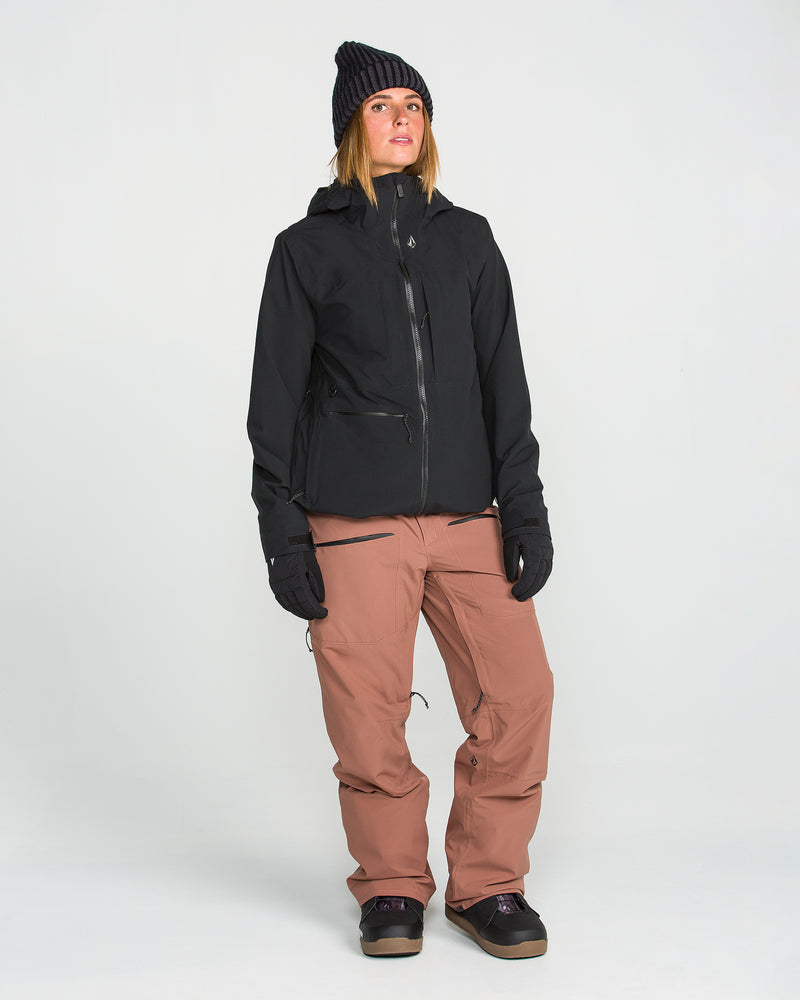 A person is seen wearing the Volcom Women’s Cloudbreaker Stretch 30K Jacket in black—a waterproof snowboard jacket made from a recycled shell—paired with black gloves, a black beanie, tan pants, and dark shoes. They stand against a plain white background, gazing slightly upward and to the side.