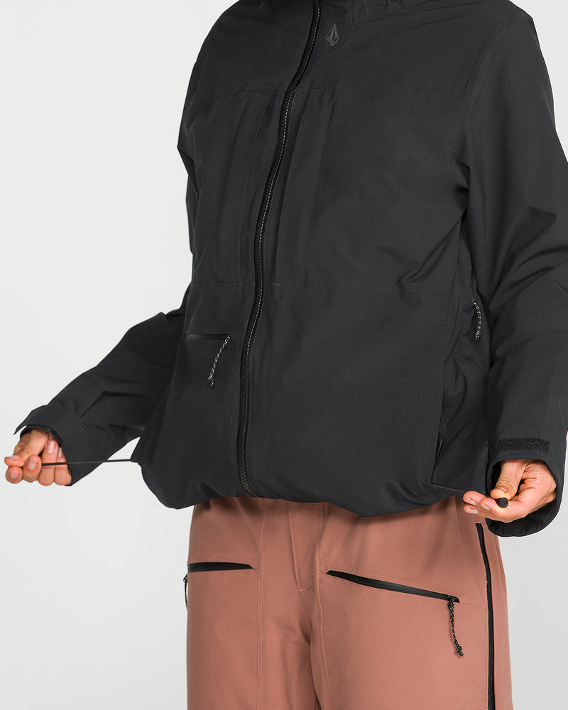 Wearing the Volcom Women’s Cloudbreaker Stretch 30K Jacket—made from recycled shell material for sustainability—in black, styled with brown pants featuring black zippers, and holding the drawstrings of the waterproof snowboard jacket at the waist; the image is cropped at the neck and knees.