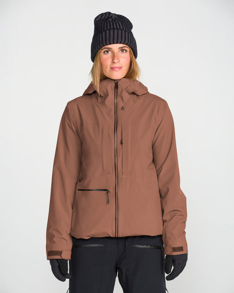 A person wearing the Volcom Women’s Cloudbreaker Stretch 30K Jacket—a waterproof jacket featuring a recycled shell—along with black gloves, black pants, and a dark knit beanie, stands facing forward against a plain light background.