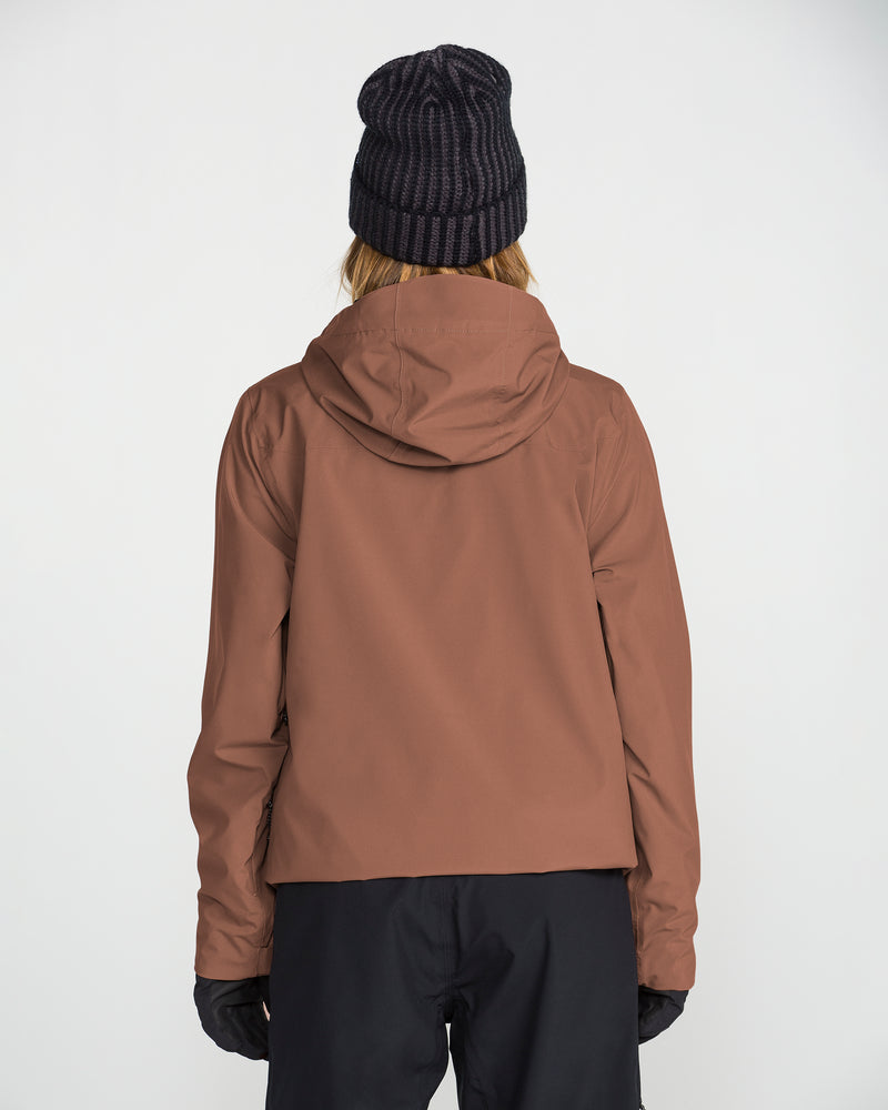 A woman stands facing away against a plain white background, wearing the Volcom Women’s Cloudbreaker Stretch 30K Jacket in brown—crafted with a recycled shell material—paired with a black knit beanie, black pants, and gloves.