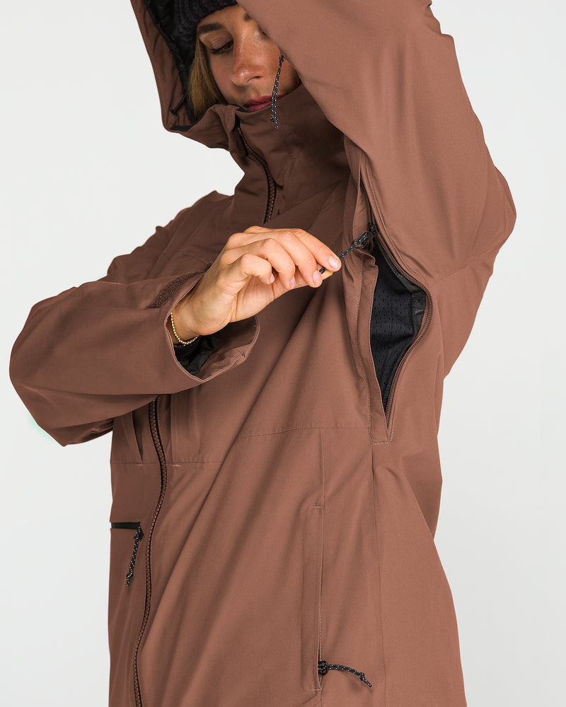 A person wearing the Volcom Women’s Cloudbreaker Stretch 30K Jacket in brown unzips an underarm ventilation opening. Only their upper body is visible, with their face partially obscured by the hood and arm; this recycled shell ski jacket delivers high-performance protection from wet conditions.