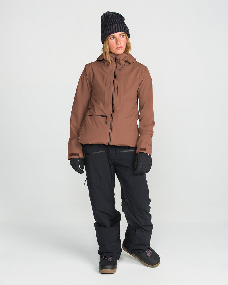 A person wearing a brown Volcom Women’s Cloudbreaker Stretch 30K Jacket, black pants, black gloves, dark shoes, and a black knit beanie stands against a plain white background.