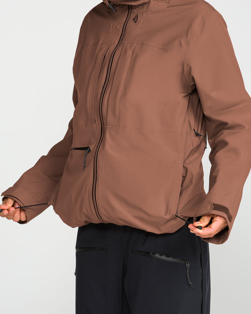 A person is adjusting the drawstrings at the hem of a brown Volcom Women’s Cloudbreaker Stretch 30K Jacket. Only their torso and hands are visible against a plain white background.