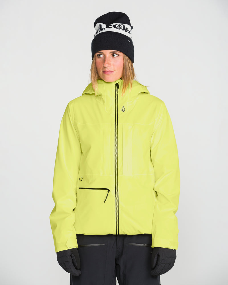 Wearing the Women’s Cloudbreaker Stretch 30K Jacket by Volcom—a bright yellow, 30K waterproof jacket crafted from a recycled shell—paired with black gloves, black pants, and a black beanie featuring white writing, this person stands against a plain white background.