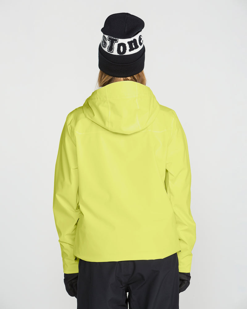 A person is facing away, wearing the Volcom Women’s Cloudbreaker Stretch 30K Jacket in bright yellow with a recycled shell, paired with black gloves, black pants, and a black beanie featuring white lettering. The background is plain white.