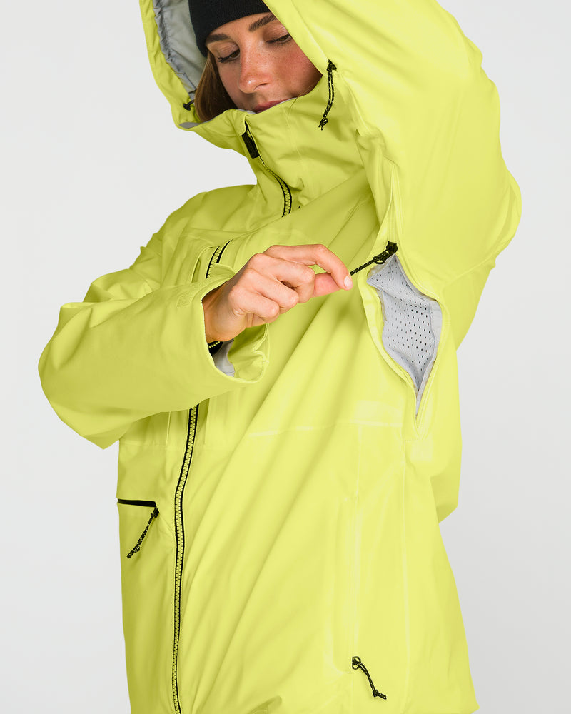 A person wearing a Volcom Women’s Cloudbreaker Stretch 30K Jacket in bright yellow unzips the underarm vent, revealing mesh lining inside. This waterproof jacket features black zippers and a hood. The person completes their look with a black beanie.