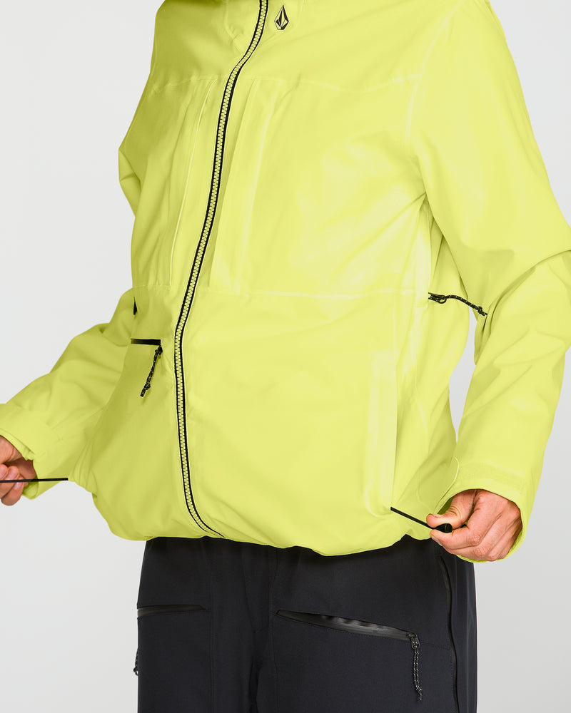 A woman wears the Volcom Women’s Cloudbreaker Stretch 30K Jacket in vibrant yellow, paired with black pants. She adjusts the drawstrings at the bottom hem of this 30K waterproof jacket, which features multiple zippered pockets and is made with a recycled shell. The background is plain and light-colored.
