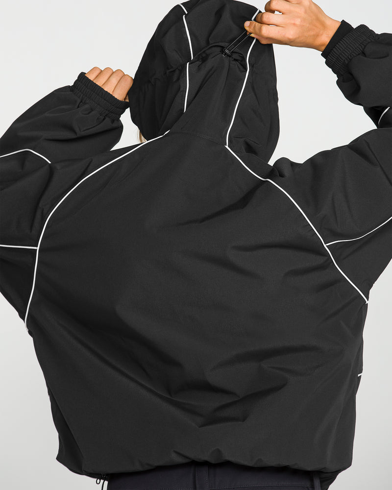 A person seen from behind wears the Volcom Unisex Liftline 20K Jacket—a black, baggy-fit snowboard jacket with white piping details—and holds the hood with both hands.