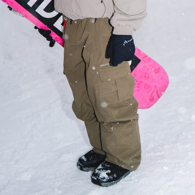 A person standing in the snow wearing Volcom cargo snow pants in a khaki brown color, paired with a beige jacket, black gloves, and black snow boots. A bright pink snowboard is visible in the background.