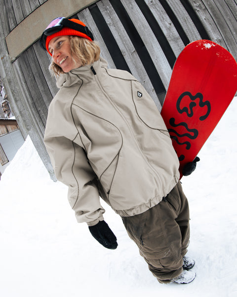 A person in winter clothes and a beanie stands in the snow, smiling and holding a red snowboard, wearing a Volcom Unisex Liftline 20K Jacket with an extra baggy fit and V-Science breathable lining, with a wooden building in the background.