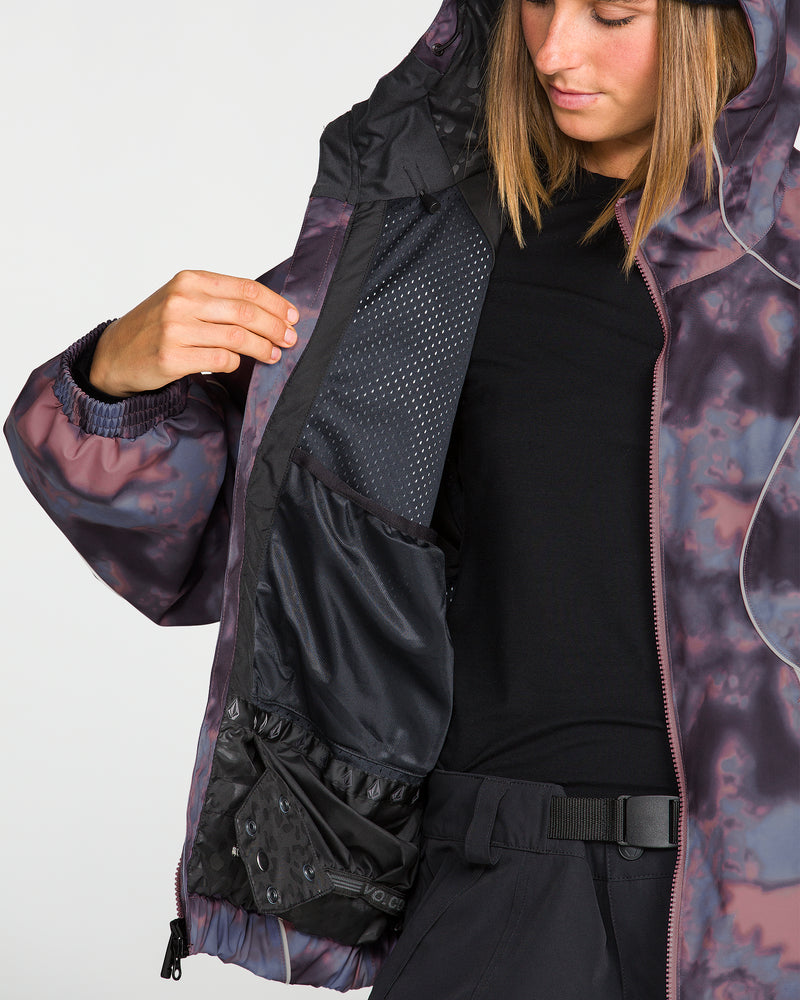 A person models the Volcom Unisex Liftline 20K Jacket, showcasing its purple patterned hooded design and baggy snowboard fit while holding it open to reveal black interior mesh pockets. This 20K waterproof jacket is styled with a black shirt and pants, with only part of their face visible.