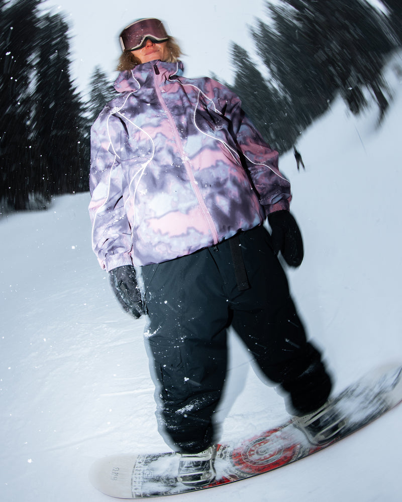 A snowboarder in a pink and purple Volcom Unisex Liftline 20K Jacket, black pants, gloves, and goggles stands on a snowboard in the snow with blurred trees in the background.