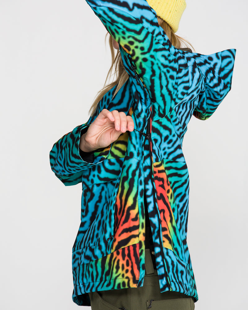 A person wearing the Volcom Women’s Harlan Pullover—a colorful, striped jacket with V-Science 2-Layer construction—and a yellow beanie raises one arm, partially obscuring their face. The bright jacket features blue, green, red, and black wavy patterns.
