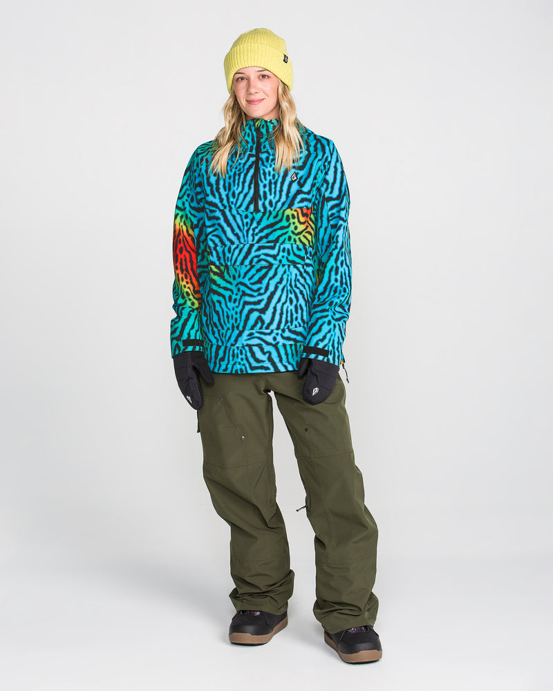 A person stands smiling, wearing a bright yellow beanie and the Volcom Women’s Harlan Pullover— a blue and black patterned jacket with red accents featuring V-Science 2-Layer fabric and PFAS-free DWR. They pair this with olive green pants, black gloves, and black boots against a plain background.