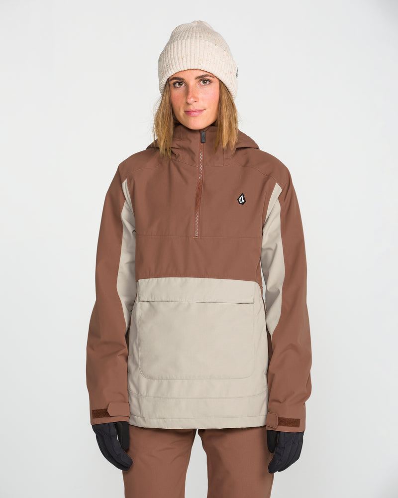 A person wearing a beige beanie, brown pants, black gloves, and the Volcom Women’s Harlan Pullover—a brown and beige jacket with a recycled shell—stands facing forward against a plain white background.
