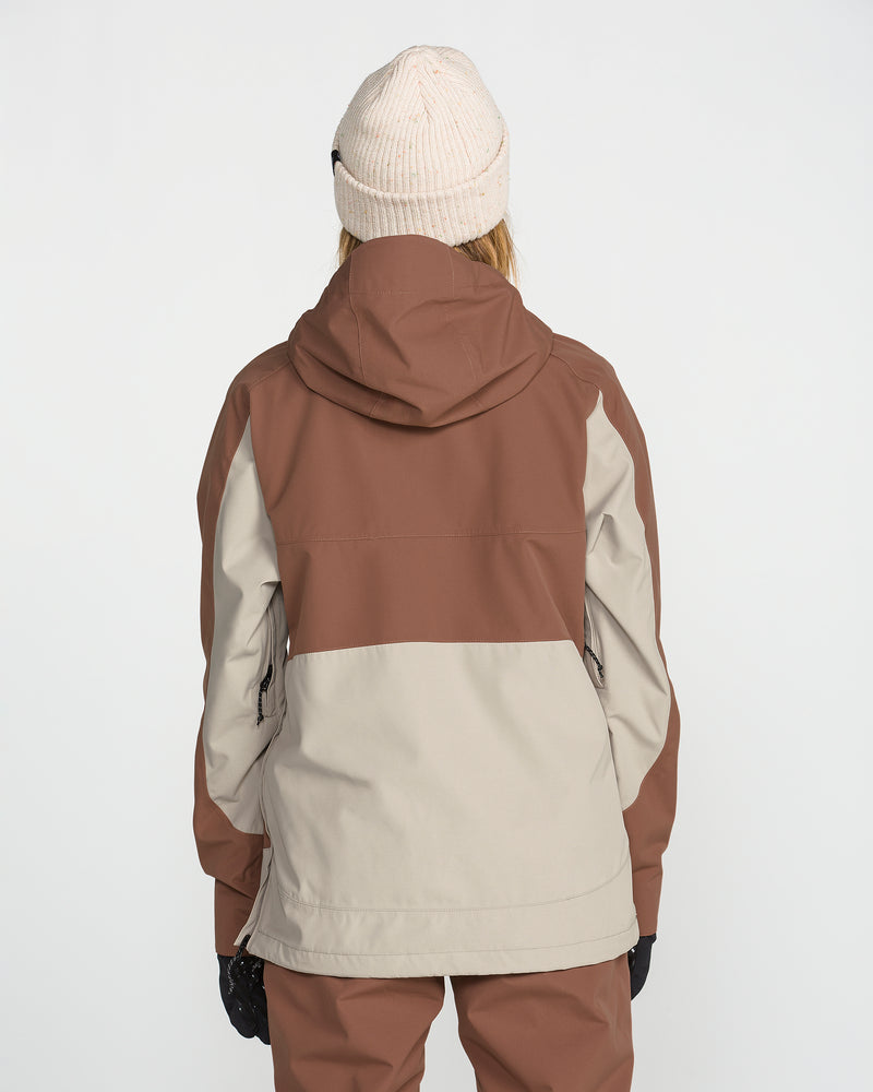 A person stands facing away from the camera against a plain white background, wearing a beige knit hat, brown pants, and the Volcom Women’s Harlan Pullover—a brown and beige waterproof jacket with a recycled shell.