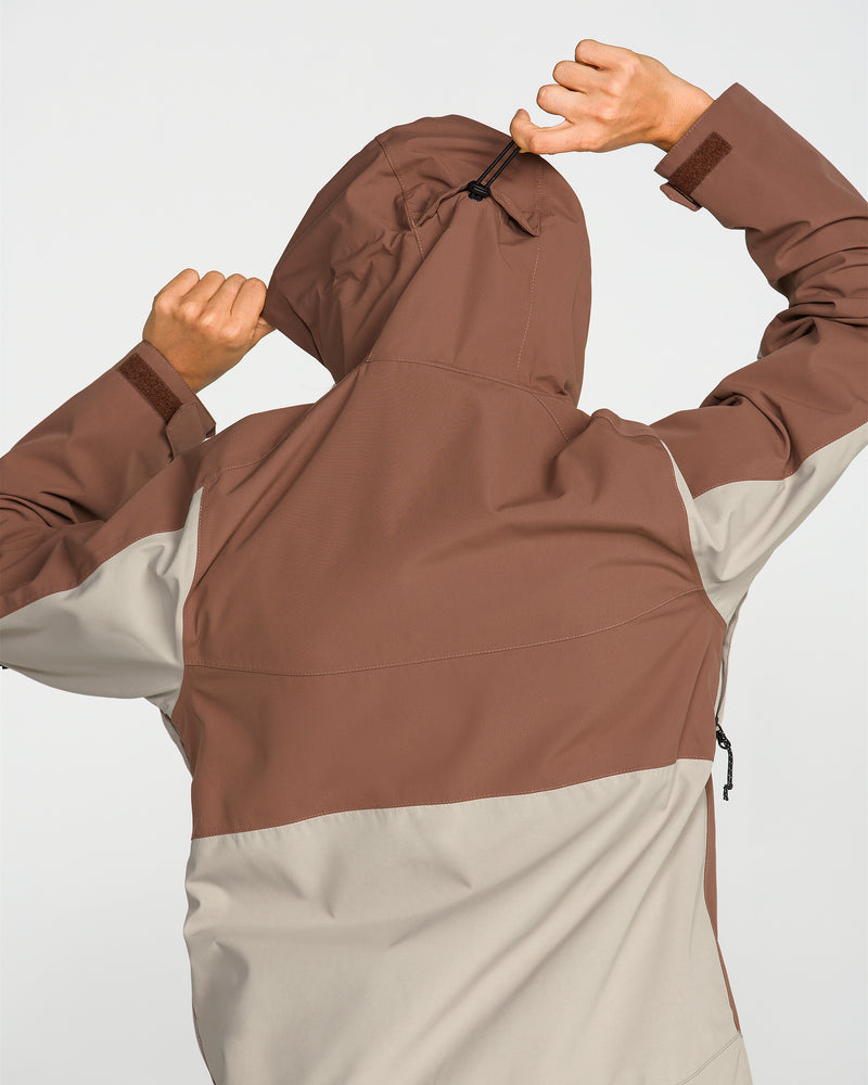 A person is seen from behind adjusting the hood of a brown and beige color-blocked waterproof jacket, identified as the Women’s Harlan Pullover by Volcom. Crafted from recycled shell material, this pullover blends practical style with sustainability; one hand pulls a drawstring while the other holds the hood’s edge, all set against a plain white background.