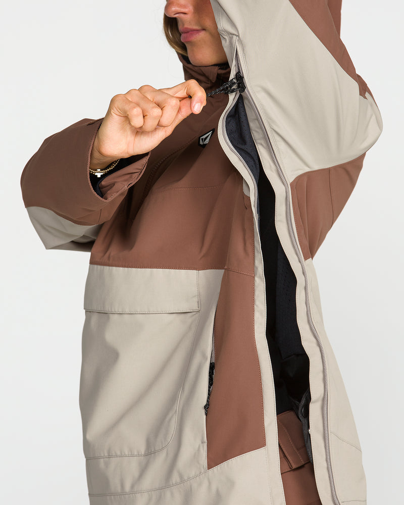 A woman wearing the Volcom Women’s Harlan Pullover in two-tone brown and beige is zipping up the front, her face partially visible as she raises one arm to adjust the hood.