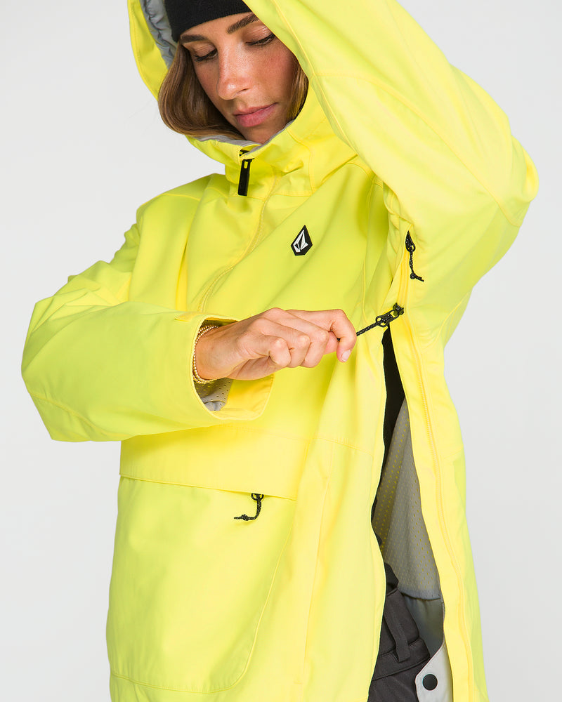 Clad in the Volcom Women’s Harlan Pullover—a bright yellow waterproof jacket featuring a black chest logo—a person unzips a side ventilation pocket. Crafted from recycled shell material, the jacket pairs effortlessly with her black beanie.