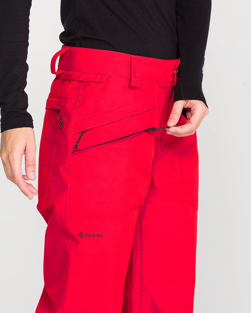 A person wearing Volcom Women’s Kanyon Insulated Gore Pants in bright red—with GORE-TEX material—and a black long-sleeve top is unzipping a pocket on the pants. Only their midsection, hand, and part of the arm are visible.