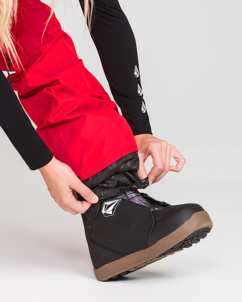 A person wearing Volcom Women’s Kanyon Insulated Gore Pants in red and a black long sleeve shirt adjusts the cuff over black and brown snowboard boots against a plain light background.