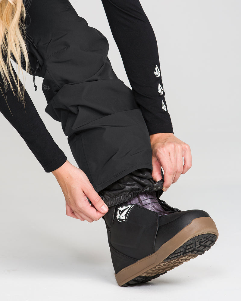 A person adjusts Volcom Women’s Kanyon Insulated Gore Pants in black over black snow boots with brown soles, both displaying a white logo. The plain, light-colored background shows only the lower body and hands.