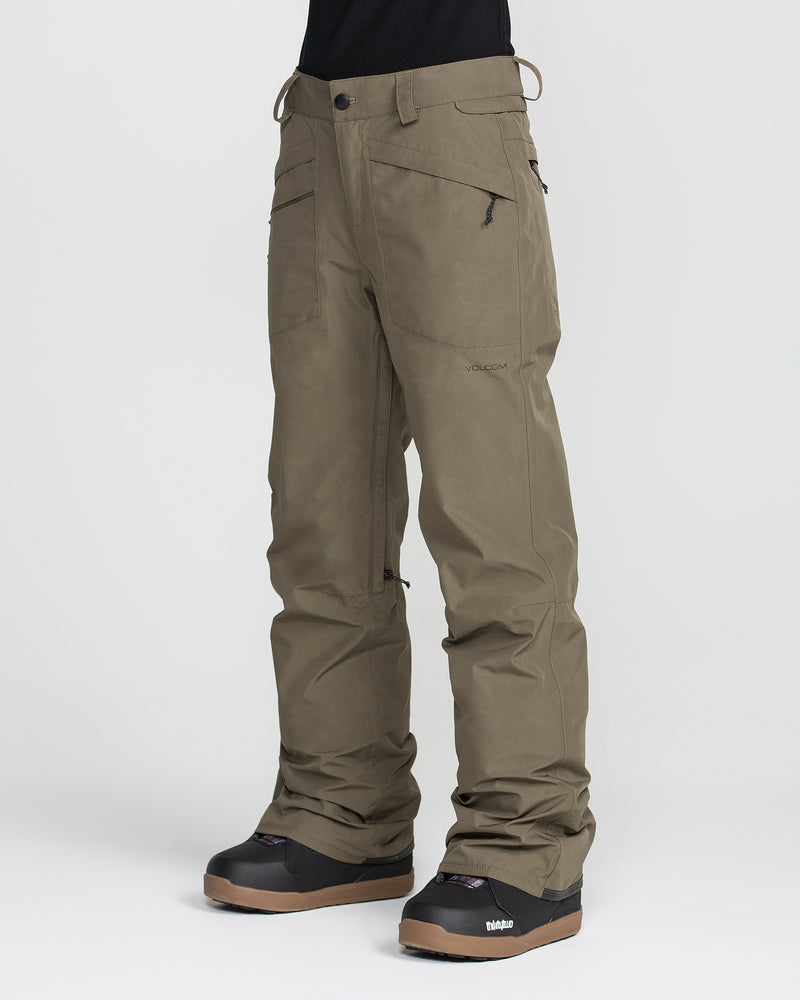 A person is shown against a plain white background wearing Volcom Women’s Kanyon Insulated Gore Pants in olive green, which are waterproof and feature multiple zippered pockets, a relaxed fit, and reinforced knees designed for outdoor or winter sports use. The outfit is paired with black snow boots.