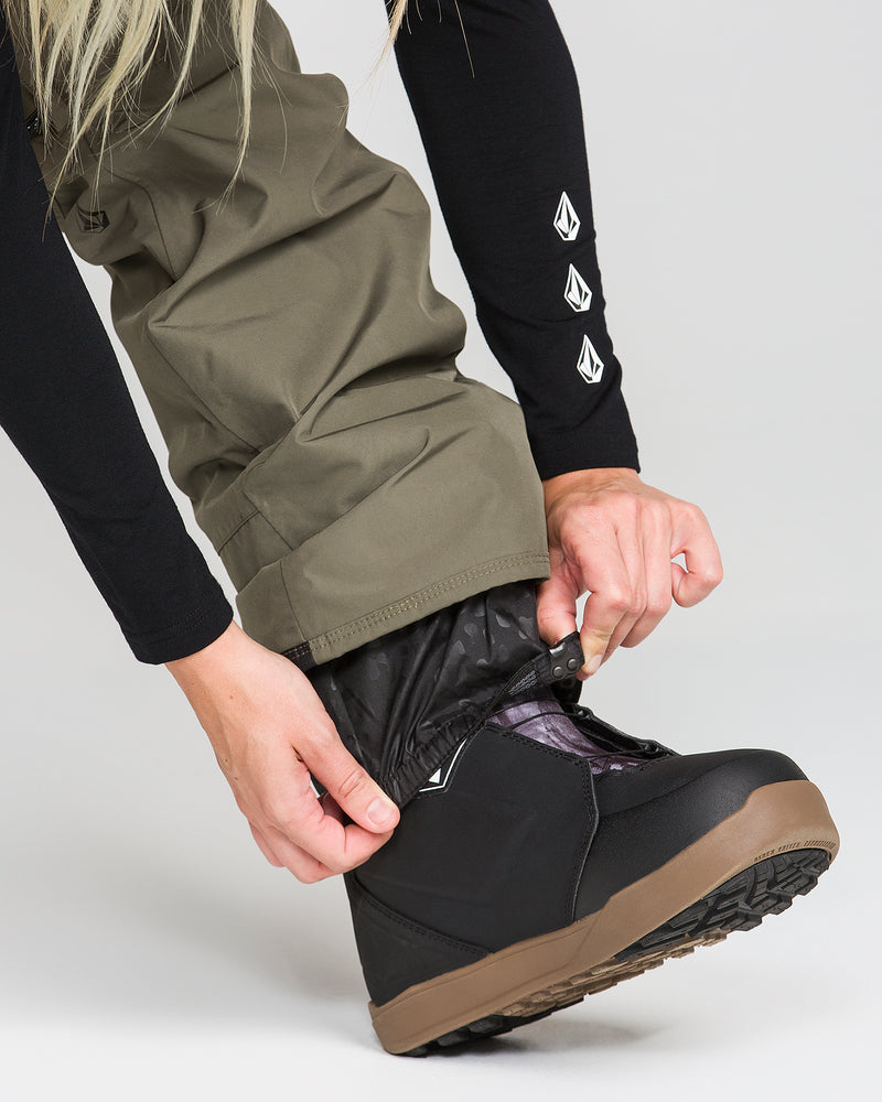 A person adjusts the olive green Volcom Women’s Kanyon Insulated Gore Pants over a black snow boot with a brown sole. Their hands secure the hem, and they are wearing a black long-sleeve with white logo graphics on the sleeve, ensuring their waterproof snow pants fit snugly for winter comfort.