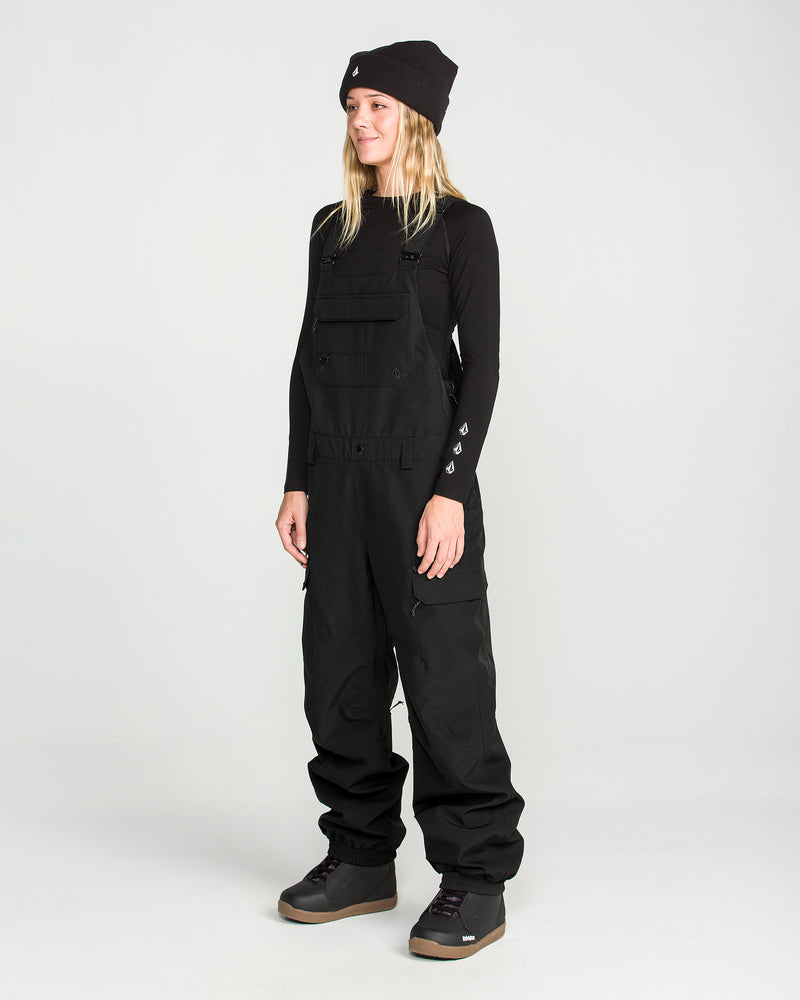 Wearing Volcom’s Women’s Creston 3D Stretch Bib made from recycled stretch oxford fabric, a black long-sleeve shirt, black beanie, and gray boots, a person stands against a plain light background, facing slightly to the left and smiling.