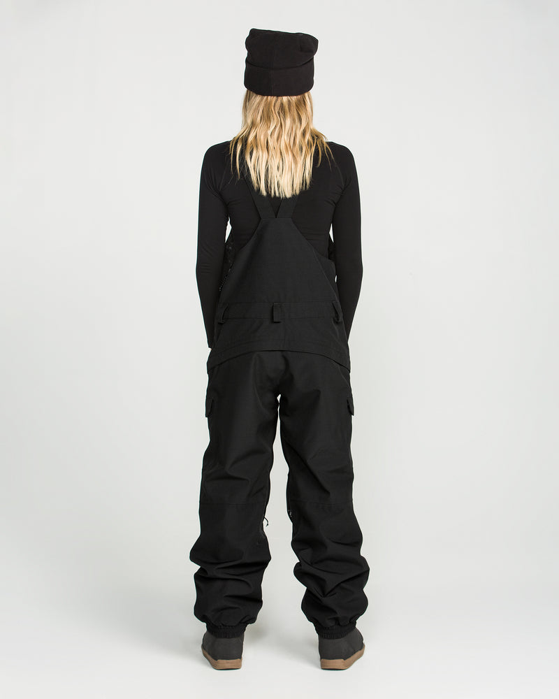 A person with long blonde hair, wearing a black beanie, a black long-sleeve shirt, and Volcom Women’s Creston 3D Stretch Bib overalls—crafted from recycled stretch oxford fabric—stands facing away against a plain white background.
