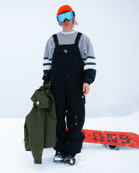 A person stands in the snow wearing Volcom’s Women’s Creston 3D Stretch Bib—black snow overalls with fully taped seams—and a striped shirt, holding a green jacket. They complete their look with a red helmet, blue goggles, and have a red snowboard on the ground beside them.