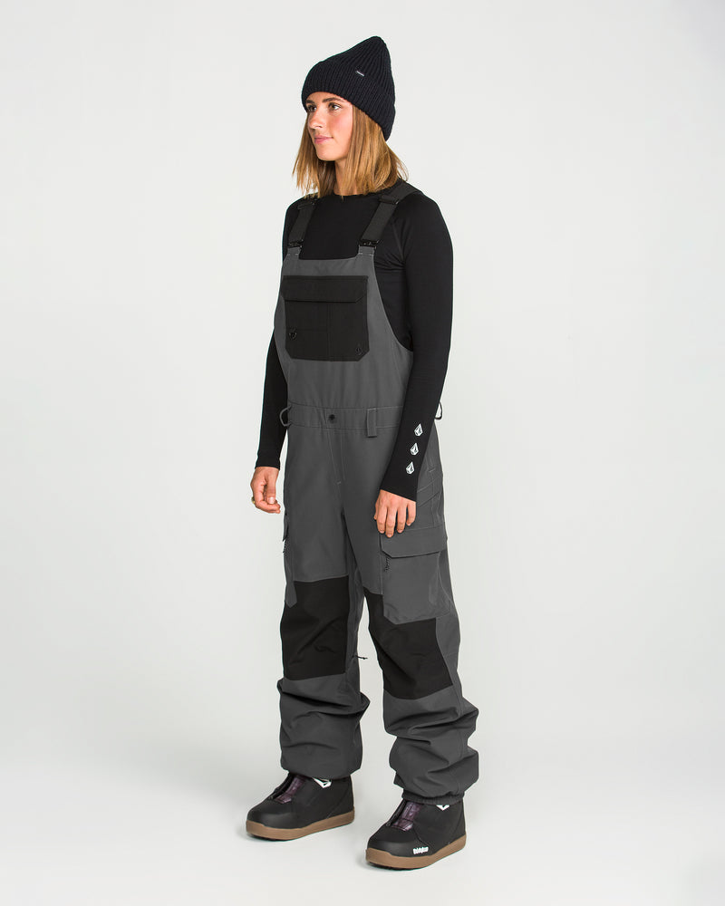 A person wearing a black beanie, a black long-sleeve shirt, and Volcom Women’s Creston 3D Stretch Bib overalls in gray with fully taped seams and black patches stands facing left against a plain white background.