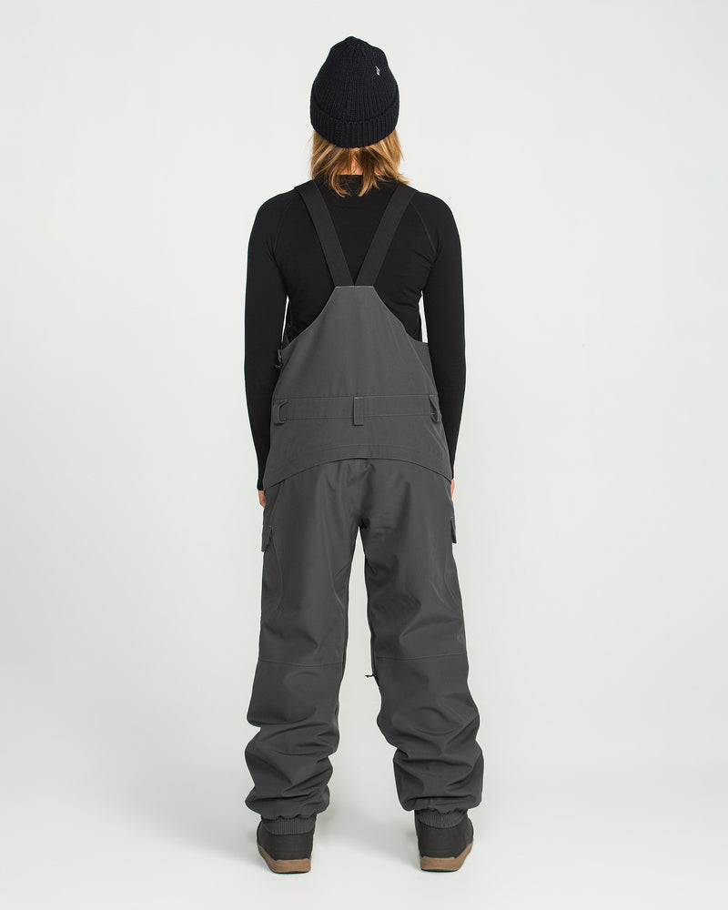 A woman stands facing away, dressed in a black beanie, black long-sleeve shirt, and Volcom Women’s Creston 3D Stretch Bib overalls in gray with wide legs and suspenders. The bib is crafted from recycled stretch oxford with fully taped seams, set against a plain white background.