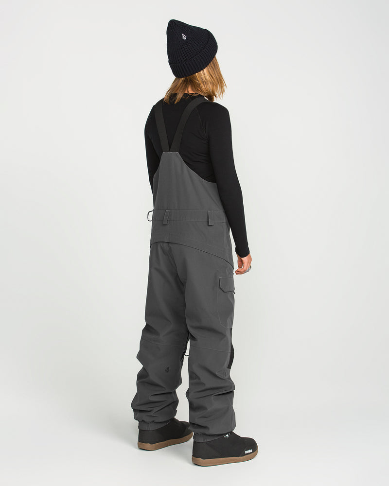 A person stands facing away, dressed in a black beanie, long-sleeve black shirt, and the Volcom Women’s Creston 3D Stretch Bib—crafted from recycled stretch oxford fabric with fully taped seams—in gray, paired with black boots against a plain white background.