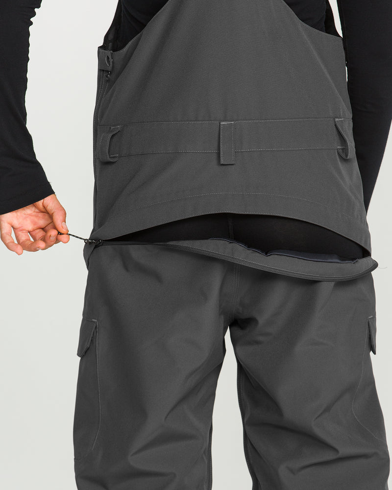 A person wearing the Volcom Women’s Creston 3D Stretch Bib in dark gray unzips a horizontal zipper at the lower back, revealing an underlayer. The image focuses on the back view, highlighting both the functional design and the recycled stretch oxford fabric with fully taped seams.