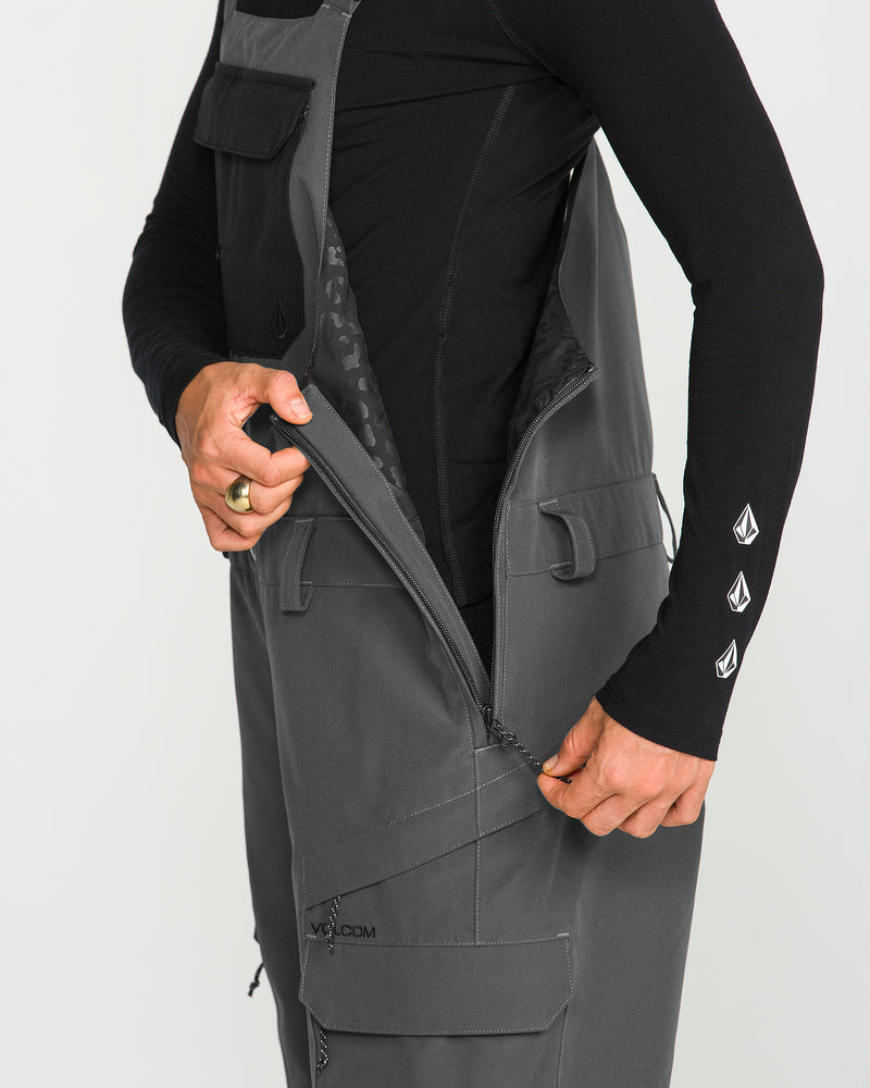 A person wearing a black long-sleeve shirt with white logos on the sleeve zips up the side of Volcom’s Women’s Creston 3D Stretch Bib in gray, showcasing its multiple pockets. The image highlights the torso and hand, drawing attention to the bib’s recycled stretch oxford fabric and fully taped seams for enhanced durability.