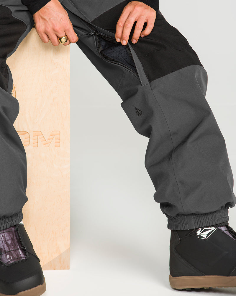 A person sits on a wooden block, unzipping the side of their gray and black Volcom Women’s Creston 3D Stretch Bib pants, crafted from recycled stretch oxford. They wear black boots with patterned detailing and a gold ring on one finger.