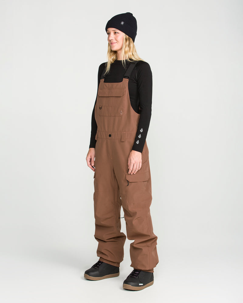 A person wearing a black beanie, a long-sleeve black shirt, and the Volcom Women’s Creston 3D Stretch Bib stands on a plain white background, looking slightly to the side with hands relaxed by their sides.