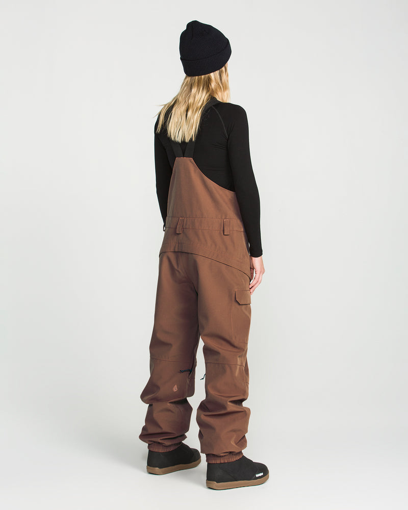 A person with long blonde hair wears the Volcom Women’s Creston 3D Stretch Bib in brown recycled stretch oxford fabric, a black beanie, and a black long-sleeve shirt. They are standing facing away from the camera against a plain white background.